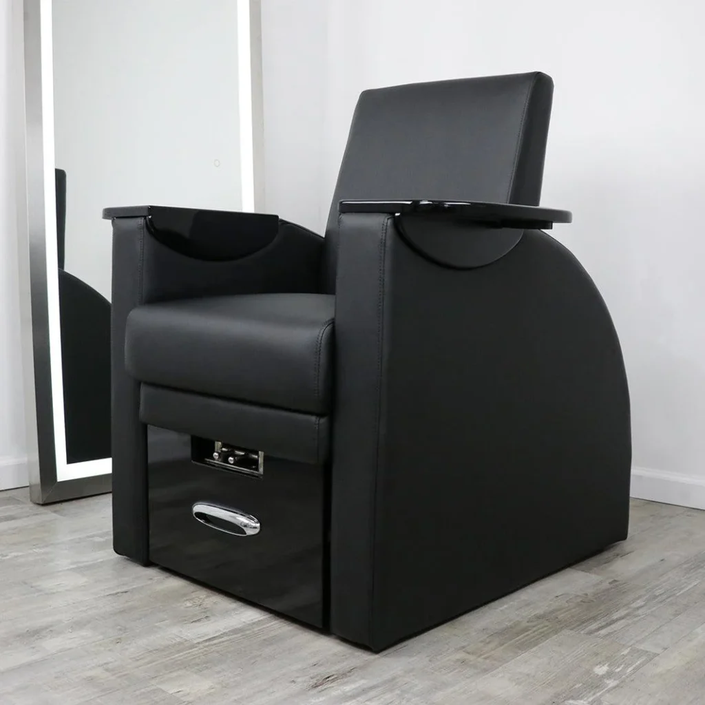 A luxurious massage pedicure chair with a high-backed design and plush, leather upholstery sits in a minimalist, well-lit studio. Warm lighting from overhead fixtures casts a soft glow, highlighting the chair's sleek, ergonomic lines. In the foreground, a metal tray holds an array of pedicure tools from the James Barber Shop, complementing the chair's elegant aesthetic. The background features a simple, light-colored wall, allowing the chair to take center stage and convey a sense of relaxation and indulgence.